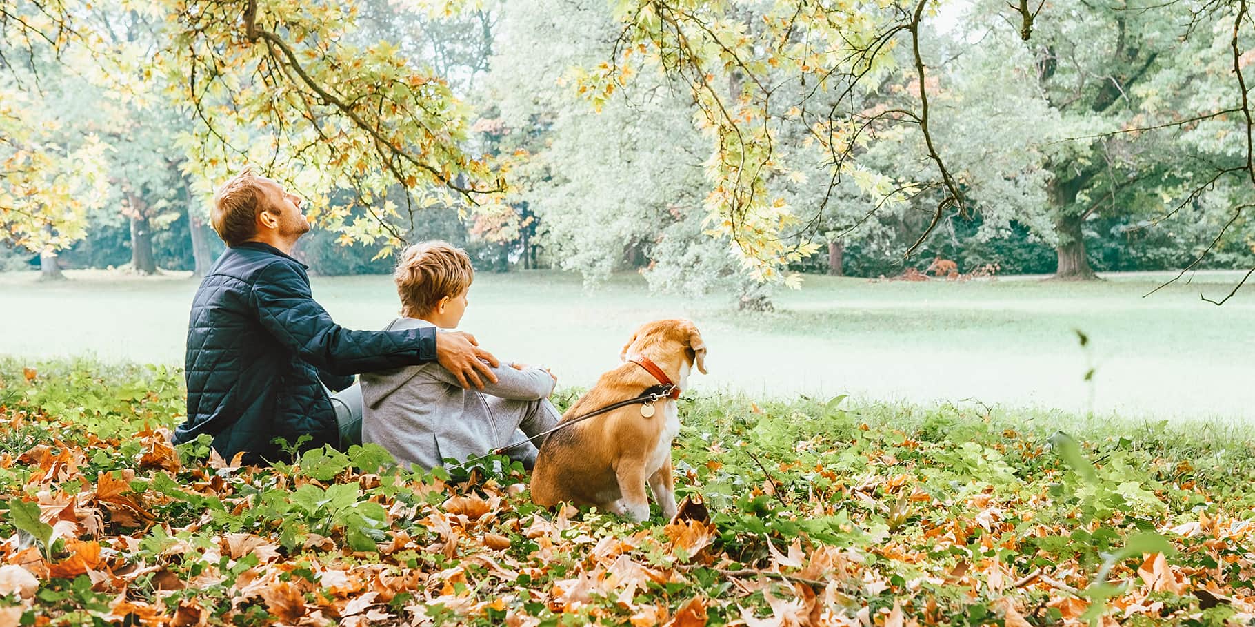 A father with his arm around his son sitting on the grass with their dog.