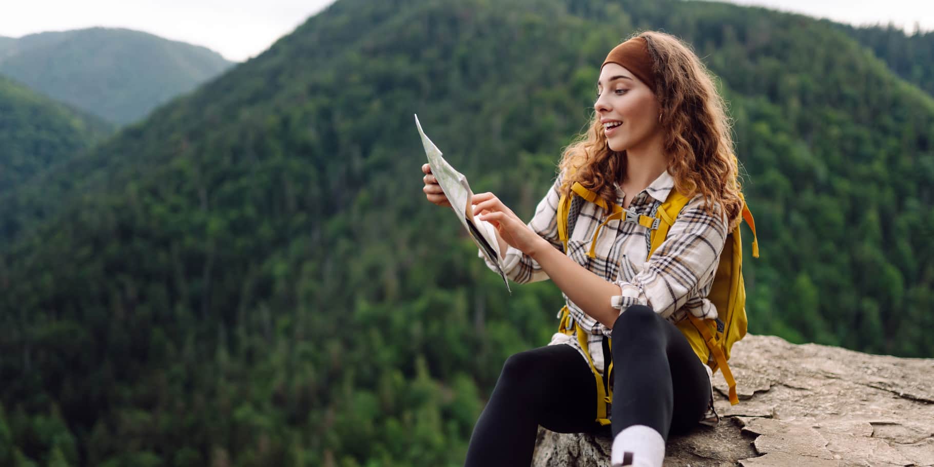 A smiling female hiker sitting on a rock looking at a map with mountains in the background.