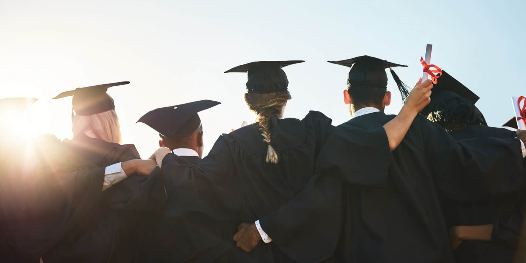 A group of graduates in caps and gowns standing arm in arm celebrating their achievement.