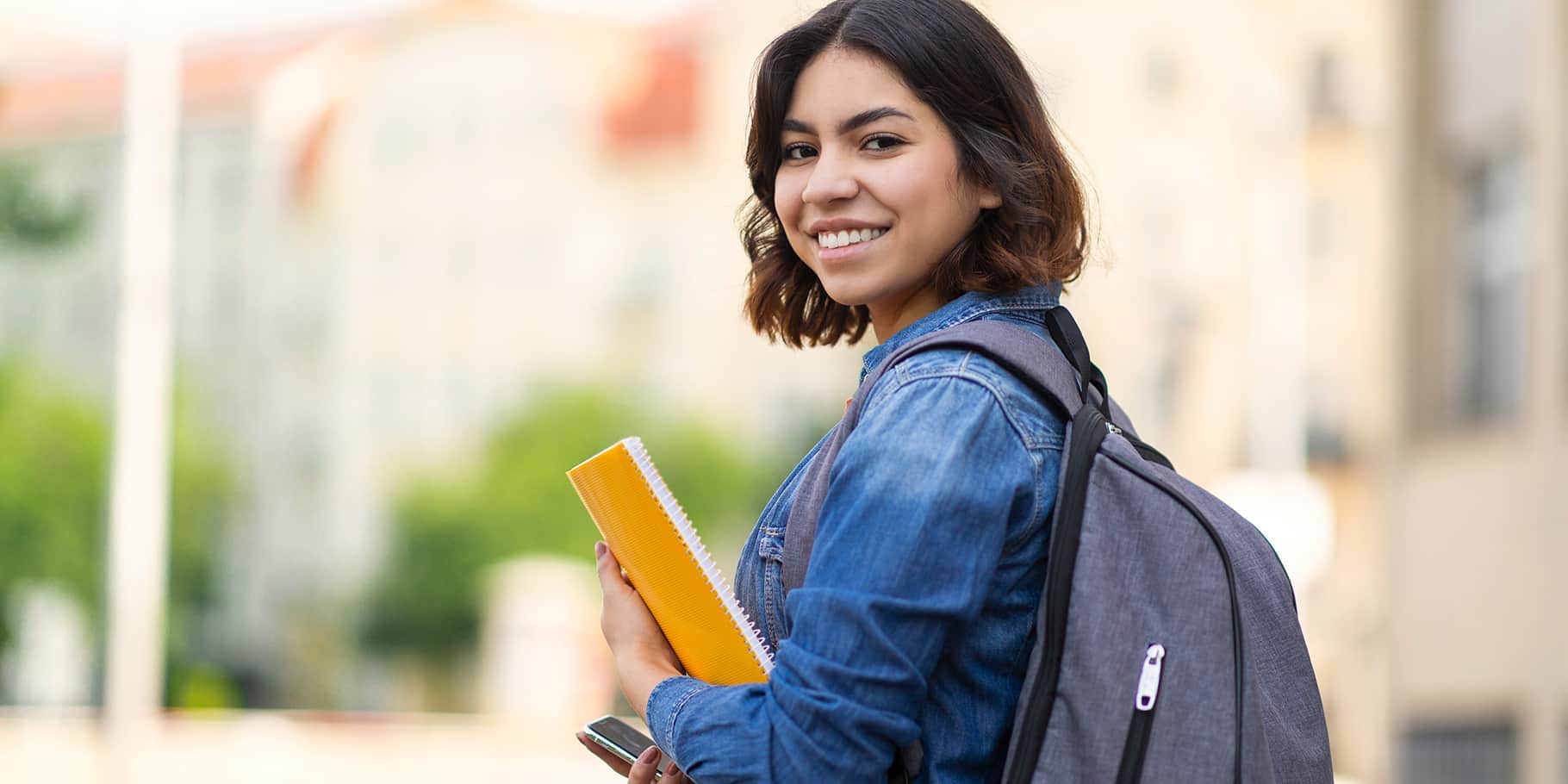 A smiling female college student carrying a bookbag and holding a notebook.